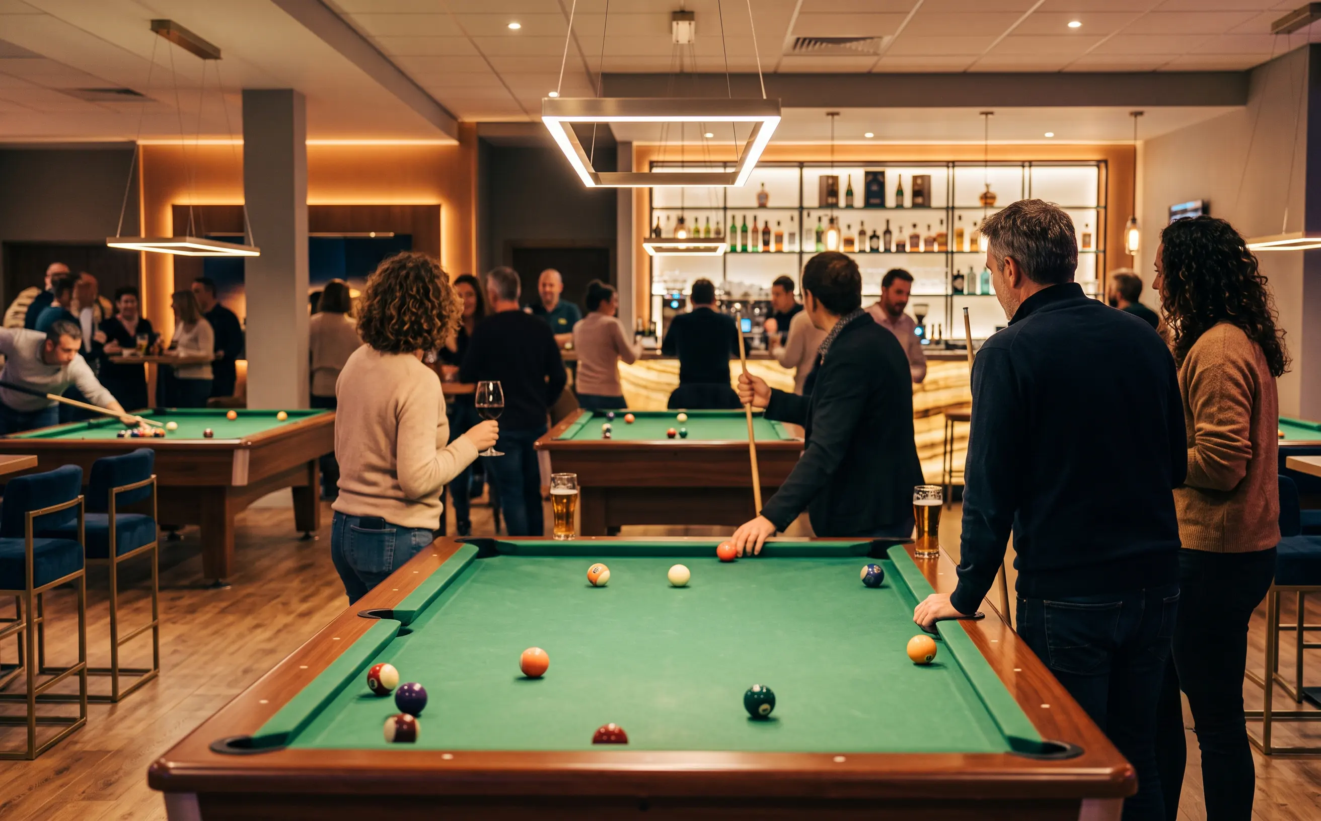 Salle de billard tamisée aux tables de tapis vert, bar en arrière-plan, autour d’une partie en cours